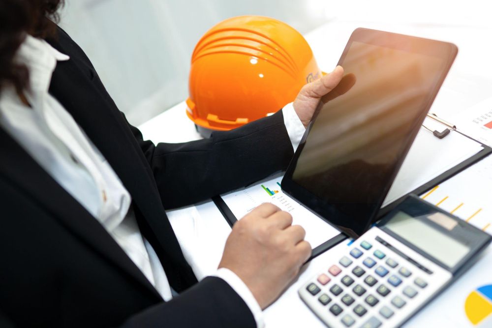 Business professional using tablet at desk with calculator and hard hat, analyzing charts and data.