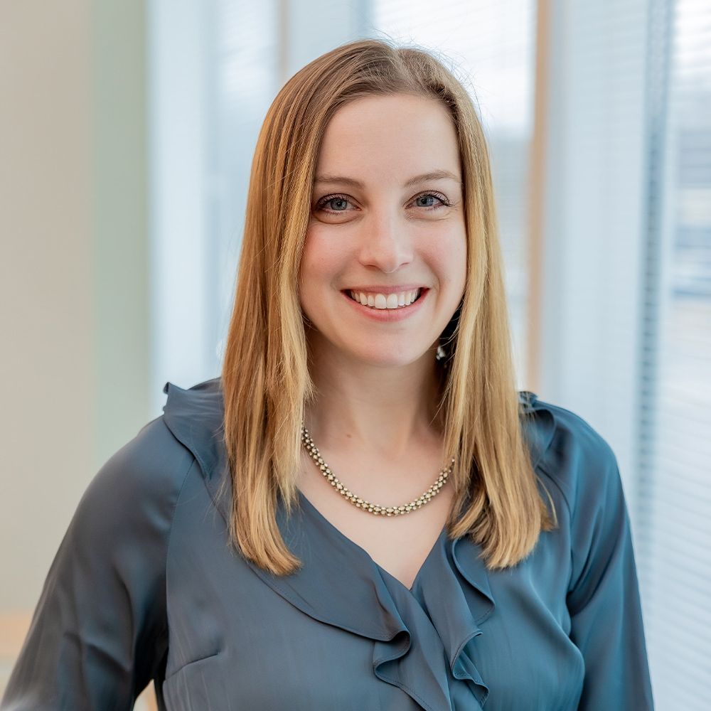 Smiling woman in blue blouse with necklace standing near window blinds.