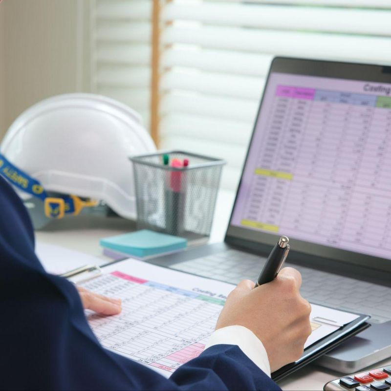 Person analyzing construction budget on laptop and clipboard with calculator and hard hat on desk.