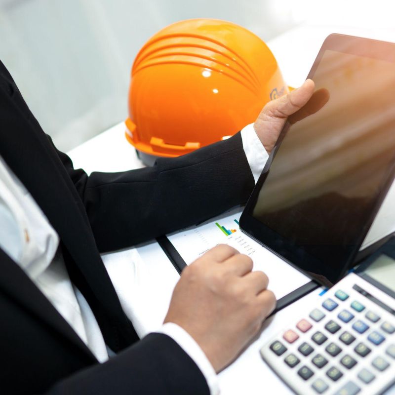 Business professional using tablet at desk with calculator and hard hat, analyzing charts and data.