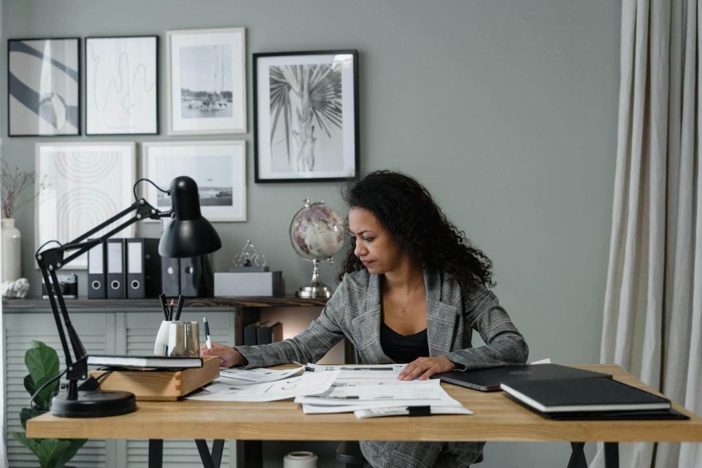 Woman in a gray suit working at a desk with papers and lamp in a stylish home office.