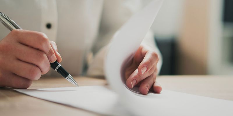 Person writing on a paper at a desk, focused on creating content or signing a document.