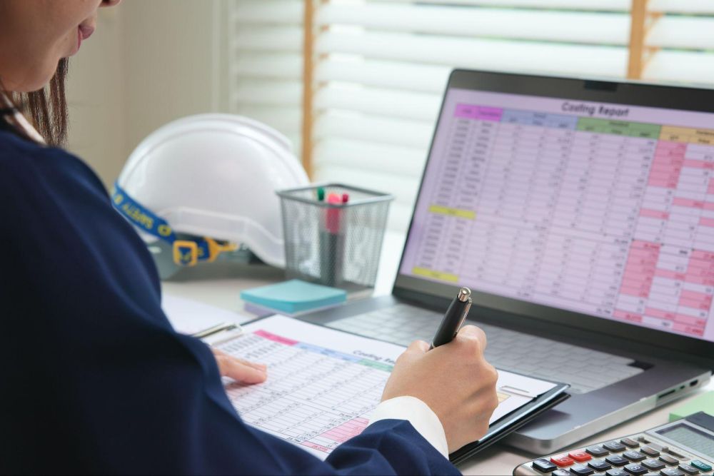 Person analyzing construction budget on laptop and clipboard with calculator and hard hat on desk.