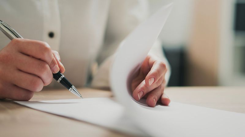 Person writing on a paper at a desk, focused on creating content or signing a document.