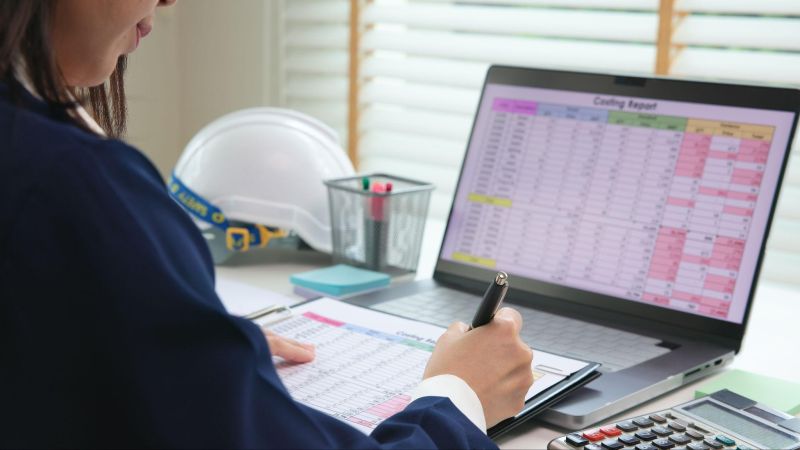Person analyzing construction budget on laptop and clipboard with calculator and hard hat on desk.