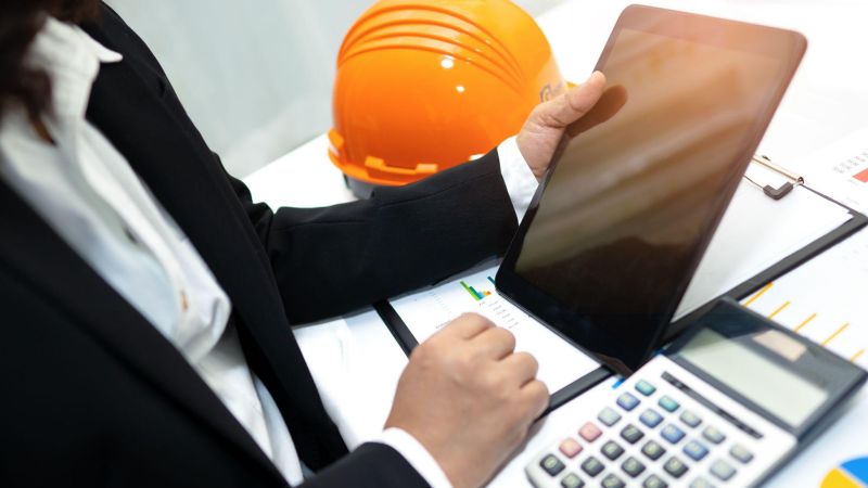 Business professional using tablet at desk with calculator and hard hat, analyzing charts and data.