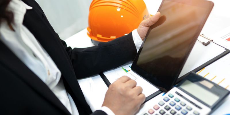 Business professional using tablet at desk with calculator and hard hat, analyzing charts and data.